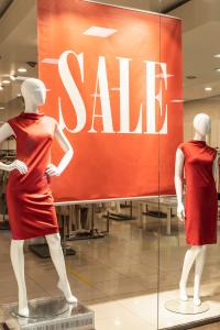 Dos maniquíes con vestidos rojos se exhiben en el escaparate de una tienda. Un gran cartel rojo con letras blancas dice "REBAJAS" al fondo, reflejando el luminoso interior de la tienda y los percheros visibles.