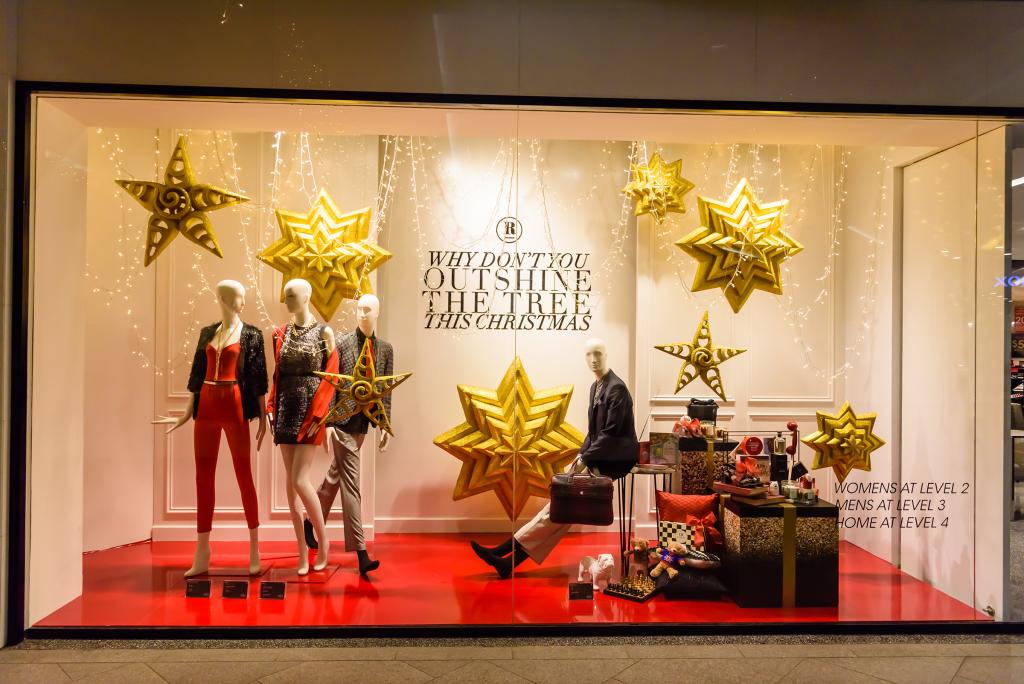 Escaparate con maniquíes vestidos con atuendos navideños, rodeados de grandes estrellas doradas. El fondo dice "¿Por qué no eclipsas el árbol esta Navidad?". Un maniquí sentado a la derecha coloca los regalos.
