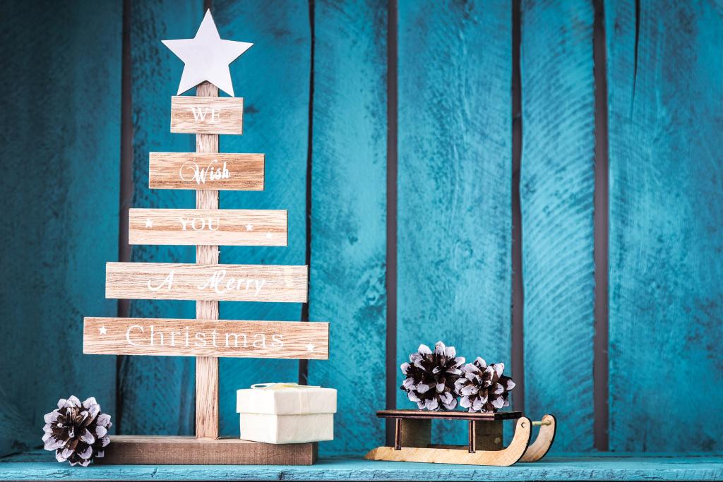 Letrero de madera para árbol de Navidad con la frase "Te deseamos una Feliz Navidad", adornado con una estrella en la parte superior. Junto a él, una pequeña caja de regalo, un trineo en miniatura y piñas. El fondo presenta tablones verticales de madera azul.